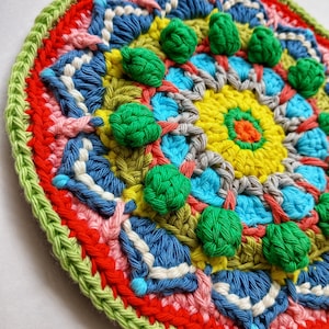 May include: A colourful crocheted mandala with a green, blue, red, pink, yellow and white colour scheme. The mandala is circular in shape and is decorated with small green crocheted balls.