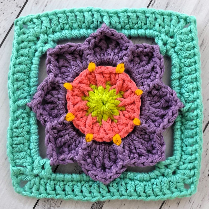 May include: A crocheted square with a purple flower in the center. The flower has a pink and yellow center and is surrounded by a teal border.