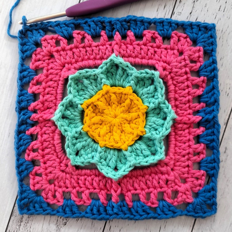 May include: A crocheted square with a multi-colored flower design. The flower is made of yellow, teal, and turquoise yarn. The square is bordered with blue and pink yarn.