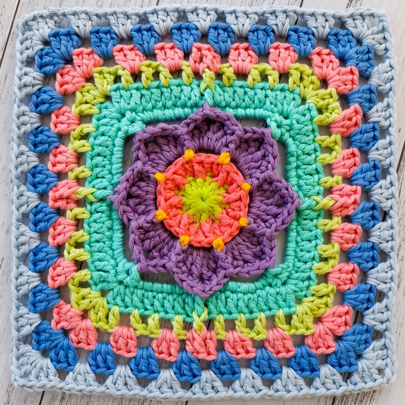 May include: A colorful crochet square with a flower design in the center. The flower is purple with a pink and green center. The square is surrounded by a border of blue, pink, and green.