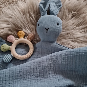 May include: A blue bunny security blanket with a textured surface and a small, multi-colored teether attached. The blanket has a button closure.