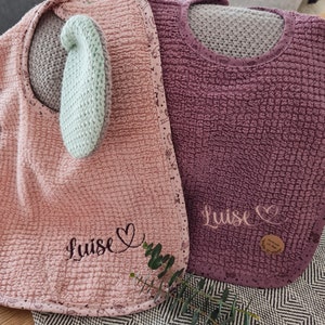 Könnte beinhalten: Zwei rosa und lila Waffel-Textur-Lätzchen mit gestickten Herzen und dem Namen "Luise" auf jedem.