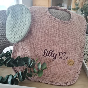 Może przedstawiać: Różowy śliniaczek dla dziecka z białą lamówką i haftowanym imieniem "Lilly" w kolorze brązowym z sercem. Śliniaczek leży na drewnianej powierzchni z zielonym wieńcem z liści.