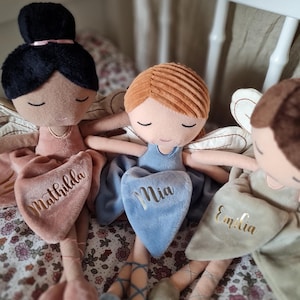 Könnte beinhalten: Drei Plüsch-Feenpuppen mit geschlossenen Augen und Ballettschuhen. Jede Puppe hat eine andere Haarfarbe und Kleiderfarbe. Die Puppen haben die gestickten Namen: "Mathilda", "Mia" und "Emilia" auf ihren herzförmigen Kleidern. Die Puppen haben Flügel.