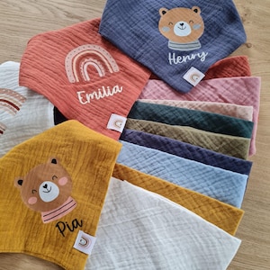 Könnte beinhalten: Ein Stapel aus zehn bunten Musselin-Babyspucktüchern mit verschiedenen Designs. Die Spucktücher zeigen einen Regenbogen, einen Bären und die Namen "Emilia", "Henry" und "Pia".