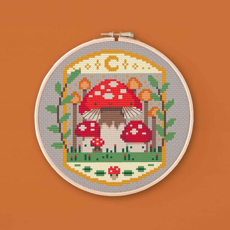 Cross stitched hoop with a toadstool design. The cross stitch design consists on a some mushrooms surrounded by a frame decorated with a moon, stars and leaves. The art style is cottagecore, witchy and whimsical. The colors of the design are red, yellow, orange and green. This cross stitch design is modern, unique and original. Stitched on linen Aida fabric. Available to download as a cross PDF stitch pattern.