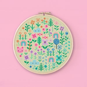 Cross stitched hoop with a kawaii design. The cross stitch design consists on flowers, leaves, rainbows, candy, clouds and a pink bear arranged in a round shape. The art style is cute and kawaii. The colors of the design are pastel, mainly orange, pink, purple, blue and teal. This cross stitch design is modern, minimalist and colorful. Stitched on light yellow Aida fabric. Available to download as a cross PDF stitch pattern.