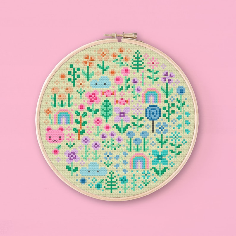 Cross stitched hoop with a kawaii design. The cross stitch design consists on flowers, leaves, rainbows, candy, clouds and a pink bear arranged in a round shape. The art style is cute and kawaii. The colors of the design are pastel, mainly orange, pink, purple, blue and teal. This cross stitch design is modern, minimalist and colorful. Stitched on light yellow Aida fabric. Available to download as a cross PDF stitch pattern.