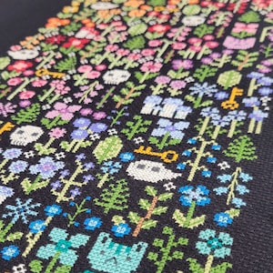 Close up of the embroidered Rainbow Garden. There are many colorful flowers, cats and mushrooms arranged by the colors of the rainbow. There are also green leaves and braches and some ecru ghosts and skulls. Stitched on black Aida. Modern and vibrant