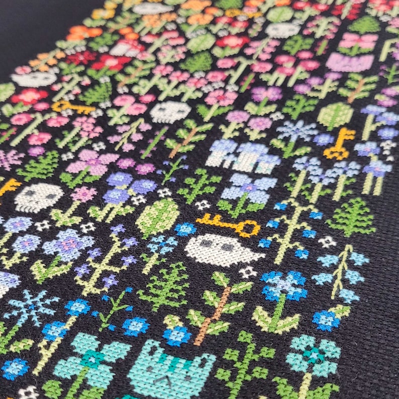 Close up of the embroidered Rainbow Garden. There are many colorful flowers, cats and mushrooms arranged by the colors of the rainbow. There are also green leaves and braches and some ecru ghosts and skulls. Stitched on black Aida. Modern and vibrant