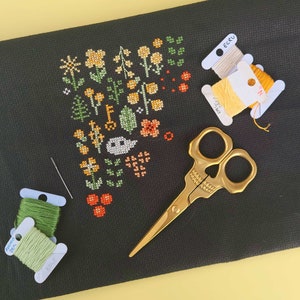 Rainbow Garden project in progress. A piece of black Aida with some yellow and orange flowers, some green leaves and an ecru ghost. Around the embroidery, there are a skull-shaped pair of scissors, a needle and some yellow and green floss bobbins.