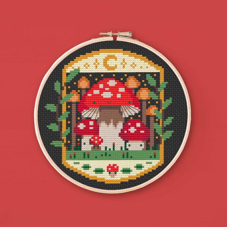 Cross stitched hoop with a toadstool design. The cross stitch design consists on a some mushrooms surrounded by a frame decorated with a moon, stars and leaves. The art style is cottagecore, witchy and whimsical. The colors of the design are red, yellow, orange and green. This cross stitch design is modern, unique and original. Stitched on black Aida fabric. Available to download as a cross PDF stitch pattern.