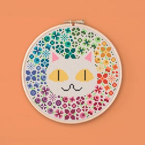 May include: White cat face cross stitch pattern with a colorful floral wreath around the face. The cat has yellow eyes and a pink nose.