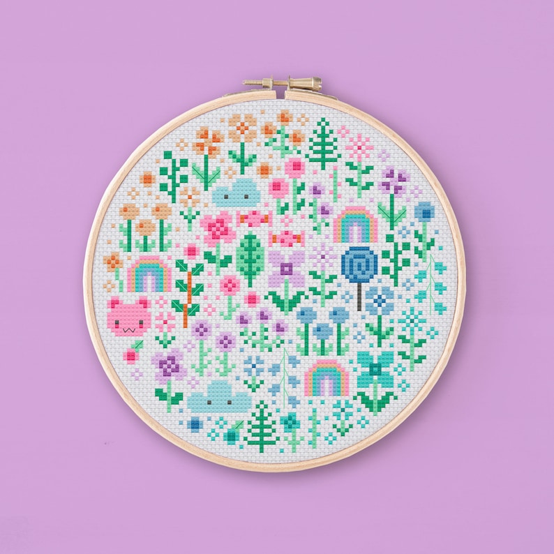 Cross stitched hoop with a kawaii design. The cross stitch design consists on flowers, leaves, rainbows, candy, clouds and a pink bear arranged in a round shape. The art style is cute and kawaii. The colors of the design are pastel, mainly orange, pink, purple, blue and teal. This cross stitch design is modern, minimalist and colorful. Stitched on white Aida fabric. Available to download as a cross PDF stitch pattern.
