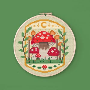 Cross stitched hoop with a toadstool design. The cross stitch design consists on a some mushrooms surrounded by a frame decorated with a moon, stars and leaves. The art style is cottagecore, witchy and whimsical. The colors of the design are red, yellow, orange and green. This cross stitch design is modern, unique and original. Stitched on light yellow Aida fabric. Available to download as a cross PDF stitch pattern.