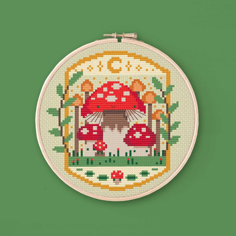 Cross stitched hoop with a toadstool design. The cross stitch design consists on a some mushrooms surrounded by a frame decorated with a moon, stars and leaves. The art style is cottagecore, witchy and whimsical. The colors of the design are red, yellow, orange and green. This cross stitch design is modern, unique and original. Stitched on light yellow Aida fabric. Available to download as a cross PDF stitch pattern.