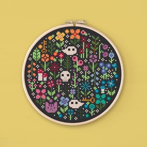 May include: A cross-stitch embroidery hoop with a floral design incorporating skulls, keys, and a cat. The flowers are in various colours, including orange, yellow, pink, blue, and purple, set against a black backdrop. The hoop is made of wood.