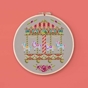 Puede incluir: Bordado de punto de cruz de un carrusel en un aro de madera. El carrusel tiene cuatro caballos y un techo a rayas rojas y blancas con detalles coloridos. Los caballos son azules, morados y amarillos. Un diseño floral en la parte inferior.