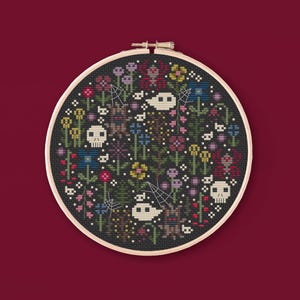 May include: A circular wooden embroidery hoop containing a cross-stitch pattern on black fabric. The design features white skulls, ghosts, red spiders, spiderwebs, and various colorful flowers in shades of pink, blue, yellow, and green.