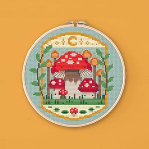 Cross stitched hoop with a toadstool design. The cross stitch design consists on a some mushrooms surrounded by a frame decorated with a moon, stars and leaves. The art style is cottagecore, witchy and whimsical. The colors of the design are red, yellow, orange and green. This cross stitch design is modern, unique and original. Stitched on sage Aida fabric. Available to download as a cross PDF stitch pattern.