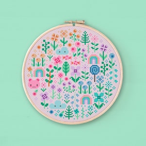 Cross stitched hoop with a kawaii design. The cross stitch design consists on flowers, leaves, rainbows, candy, clouds and a pink bear arranged in a round shape. The art style is cute and kawaii. The colors of the design are pastel, mainly orange, pink, purple, blue and teal. This cross stitch design is modern, minimalist and colorful. Stitched on light purple Aida fabric. Available to download as a cross PDF stitch pattern.