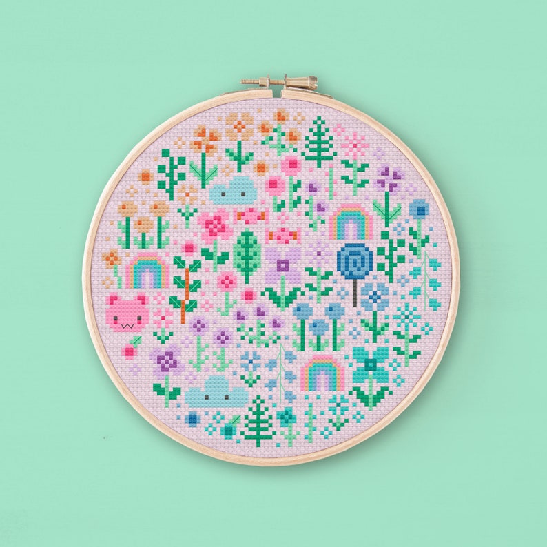 Cross stitched hoop with a kawaii design. The cross stitch design consists on flowers, leaves, rainbows, candy, clouds and a pink bear arranged in a round shape. The art style is cute and kawaii. The colors of the design are pastel, mainly orange, pink, purple, blue and teal. This cross stitch design is modern, minimalist and colorful. Stitched on light purple Aida fabric. Available to download as a cross PDF stitch pattern.