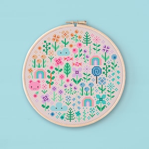 Cross stitched hoop with a kawaii design. The cross stitch design consists on flowers, leaves, rainbows, candy, clouds and a pink bear arranged in a round shape. The art style is cute and kawaii. The colors of the design are pastel, mainly orange, pink, purple, blue and teal. This cross stitch design is modern, minimalist and colorful. Stitched on light peach Aida fabric. Available to download as a cross PDF stitch pattern.