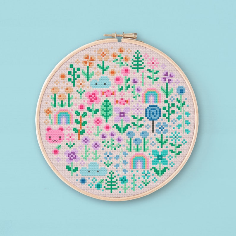 Cross stitched hoop with a kawaii design. The cross stitch design consists on flowers, leaves, rainbows, candy, clouds and a pink bear arranged in a round shape. The art style is cute and kawaii. The colors of the design are pastel, mainly orange, pink, purple, blue and teal. This cross stitch design is modern, minimalist and colorful. Stitched on light peach Aida fabric. Available to download as a cross PDF stitch pattern.