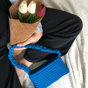Puede incluir: Una pequeña bolsa azul de crochet con una correa trenzada. La bolsa está sobre una superficie blanca y peluda.