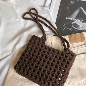 Puede incluir: Un bolso de mano de crochet marrón con forma cuadrada y un patrón tejido. El bolso tiene una correa larga y está hecho de hilo grueso.