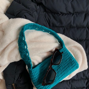 Op de afbeelding: Een turquoise gebreide handtas met een lange band en een paar zwarte zonnebrillen erin.