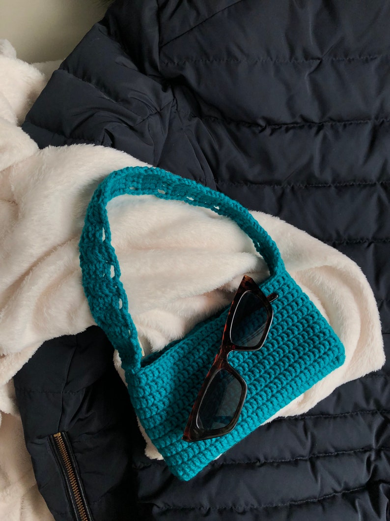 Op de afbeelding: Een turquoise gebreide handtas met een lange band en een paar zwarte zonnebrillen erin.