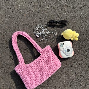 May include: A pink crocheted tote bag with two handles, alongside black sunglasses, white earbuds, a yellow plush toy, and a pink and white instant camera. The items are on a dark grey surface.