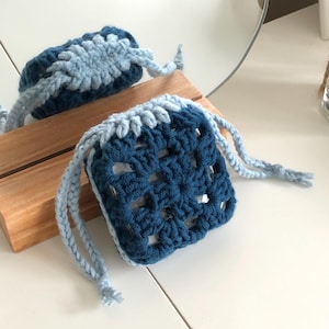 May include: A square, crocheted pouch in shades of blue. The main body is a deep navy, with a lighter blue trim and drawstring closure. The pouch is sitting on a wooden surface, with a mirror in the background.