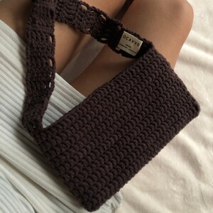 Op de afbeelding: Een donkerbruine gehaakte handtas met een lange band. De tas heeft een label met de tekst "OCAVVE SOYO HANDMADE CROCHET".