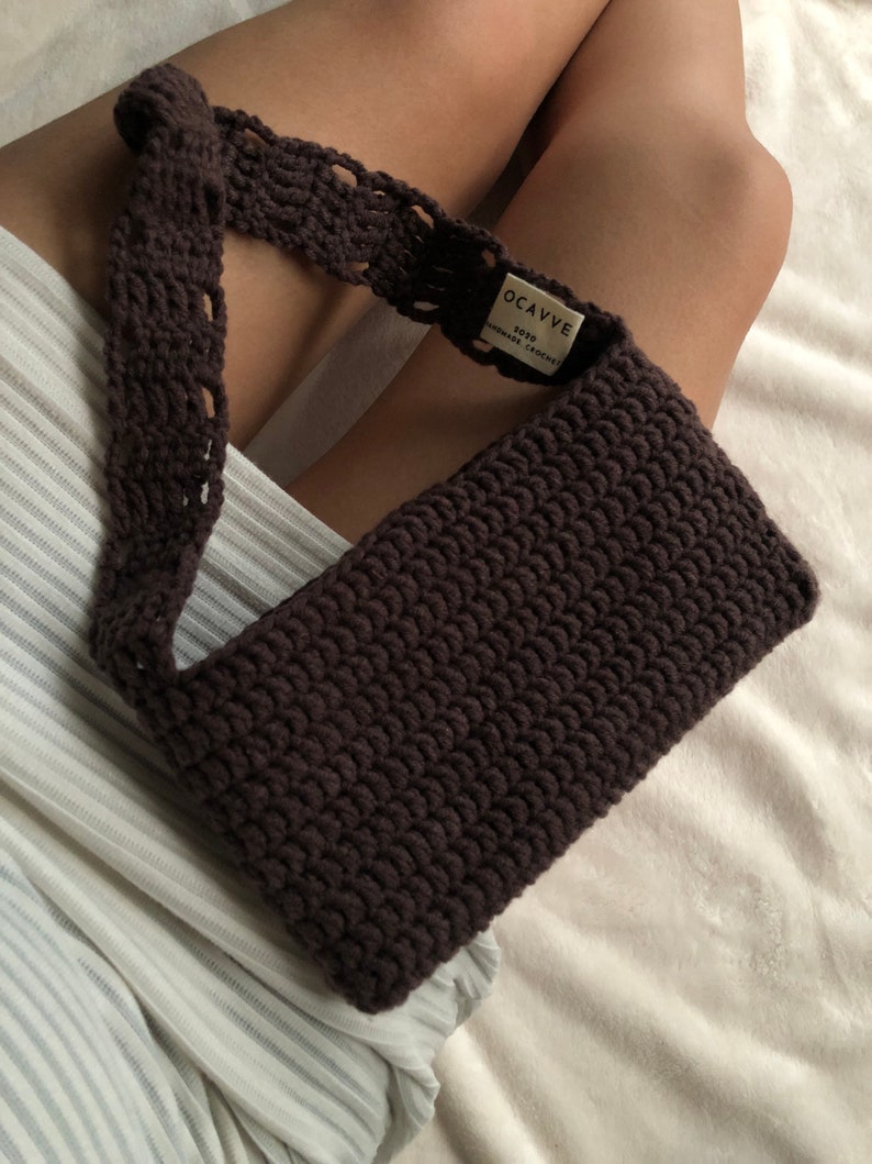 Op de afbeelding: Een donkerbruine gehaakte handtas met een lange band. De tas heeft een label met de tekst "OCAVVE SOYO HANDMADE CROCHET".