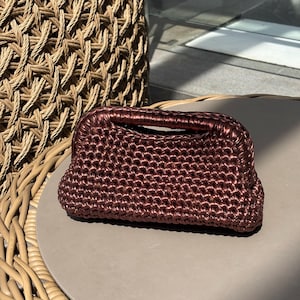 May include: A small, dark brown, woven handbag with a rounded shape and a handle. The bag is made of a textured material and sits on a neutral-colored surface. The bag is a stylish accessory.