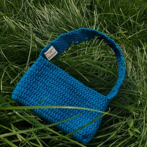 Op de afbeelding: Een felblauwe gehaakte handtas met een korte schouderband. De tas heeft een label met de tekst "OCAYVE handmade".
