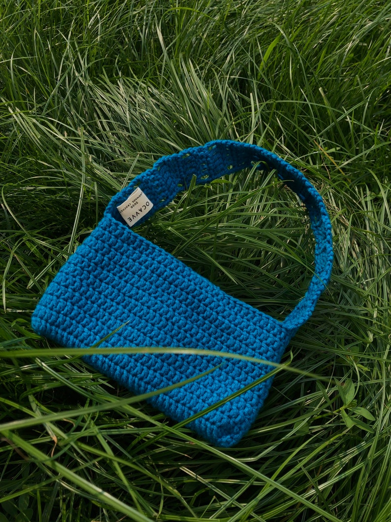 Op de afbeelding: Een felblauwe gehaakte handtas met een korte schouderband. De tas heeft een label met de tekst "OCAYVE handmade".