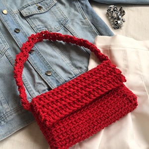 Puede incluir: Un bolso de mano de crochet rojo con una correa gruesa. El bolso está hecho de hilo grueso y tiene una forma rectangular simple.