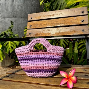 May include: A small, hand-crocheted basket with two integrated handles, featuring horizontal stripes in varying shades of purple, lavender, and mauve. The basket has a textured, thick yarn construction and a rounded base, sitting on a wooden surface.