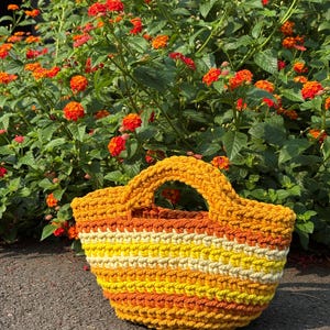 Op de afbeelding: Een handgehaakte tote bag met horizontale strepen in tinten felgeel, goudgeel en oranje. De tas is gemaakt van dik, getextureerd garen en is voorzien van twee geïntegreerde handvatten aan de bovenzijde om de tas gemakkelijk te dragen.