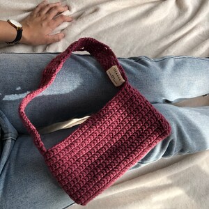 Op de afbeelding: Een bordeauxrode gehaakte handtas met een korte schouderband. De tas heeft een klein label met de tekst "OCAVVE Handmade with Love".