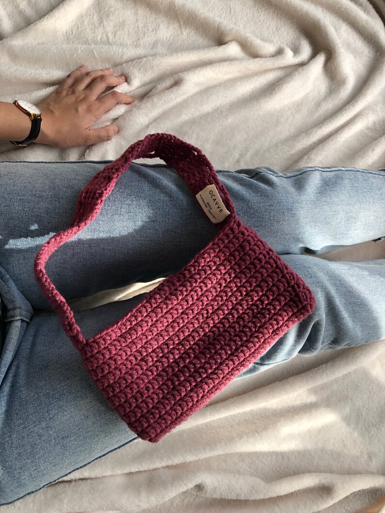 Op de afbeelding: Een bordeauxrode gehaakte handtas met een korte schouderband. De tas heeft een klein label met de tekst "OCAVVE Handmade with Love".