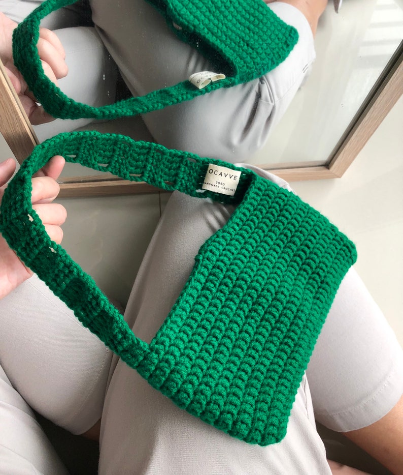 Op de afbeelding: Een groene gehaakte handtas met een klein label met de tekst "OCAVVE 2020 HANDMADE CROCHET".