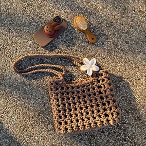 Op de afbeelding: Een bruine gehaakte tas met een open weefpatroon en een wit bloemvormig accessoire aan de schouderband. Naast de tas ligt een bruin smartphonehoesje met een klein bruin konijntje en een kleine bruine haarborstel met lichte haren.