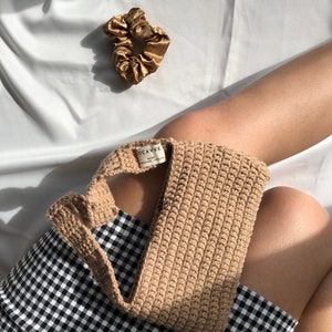 Op de afbeelding: Een bruine gehaakte tas met een zwart-wit geblokte rok op de achtergrond. De tas heeft een label met de tekst "OCAVVE 100% HANDMADE WITH LOVE".