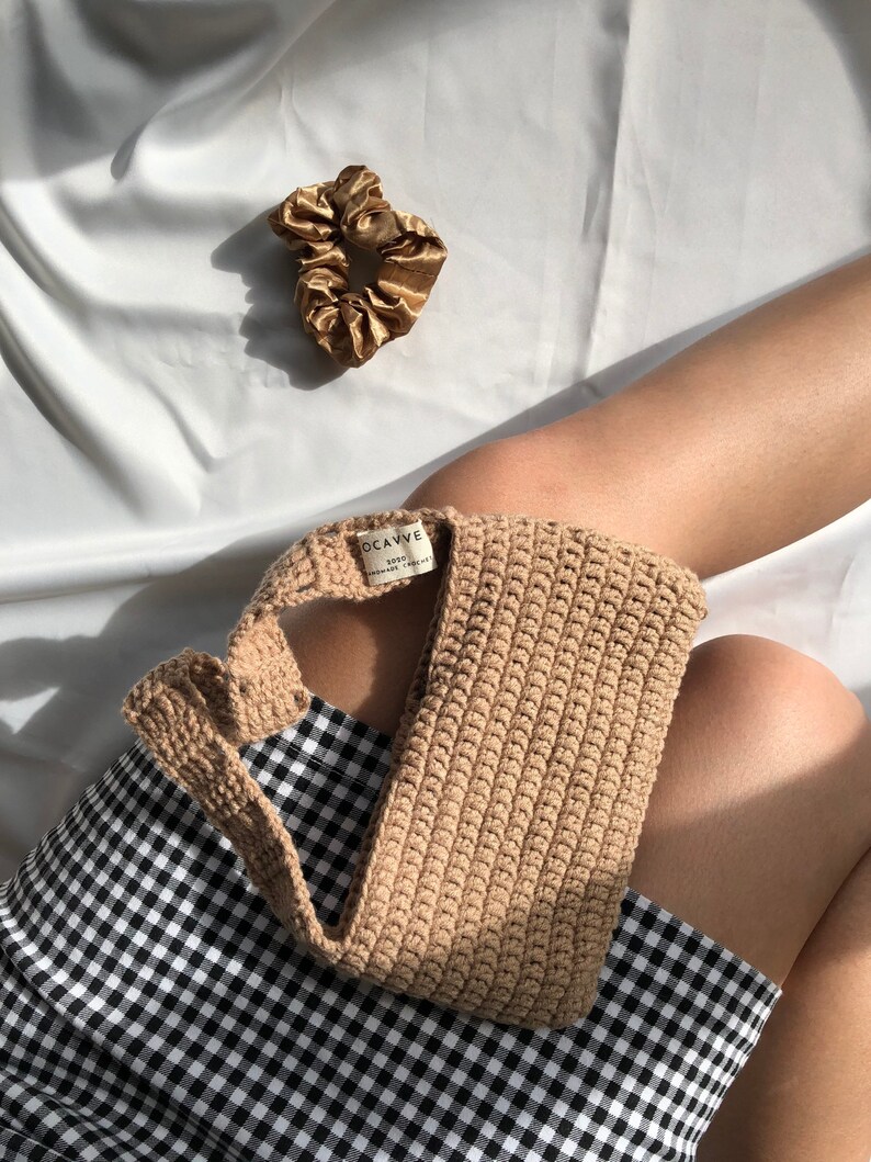Op de afbeelding: Een bruine gehaakte tas met een zwart-wit geblokte rok op de achtergrond. De tas heeft een label met de tekst "OCAVVE 100% HANDMADE WITH LOVE".