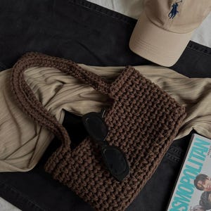 May include: A brown, crocheted handbag with a rectangular shape and a matching handle. Black sunglasses rest on top of the bag. A tan baseball cap with a blue logo and a magazine are also visible.