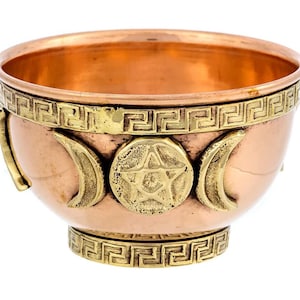 May include: A copper-colored bowl with brass accents. The bowl features a decorative band with a Greek key pattern and brass crescent moons and a star in the center. The bowl has a small handle on the side.